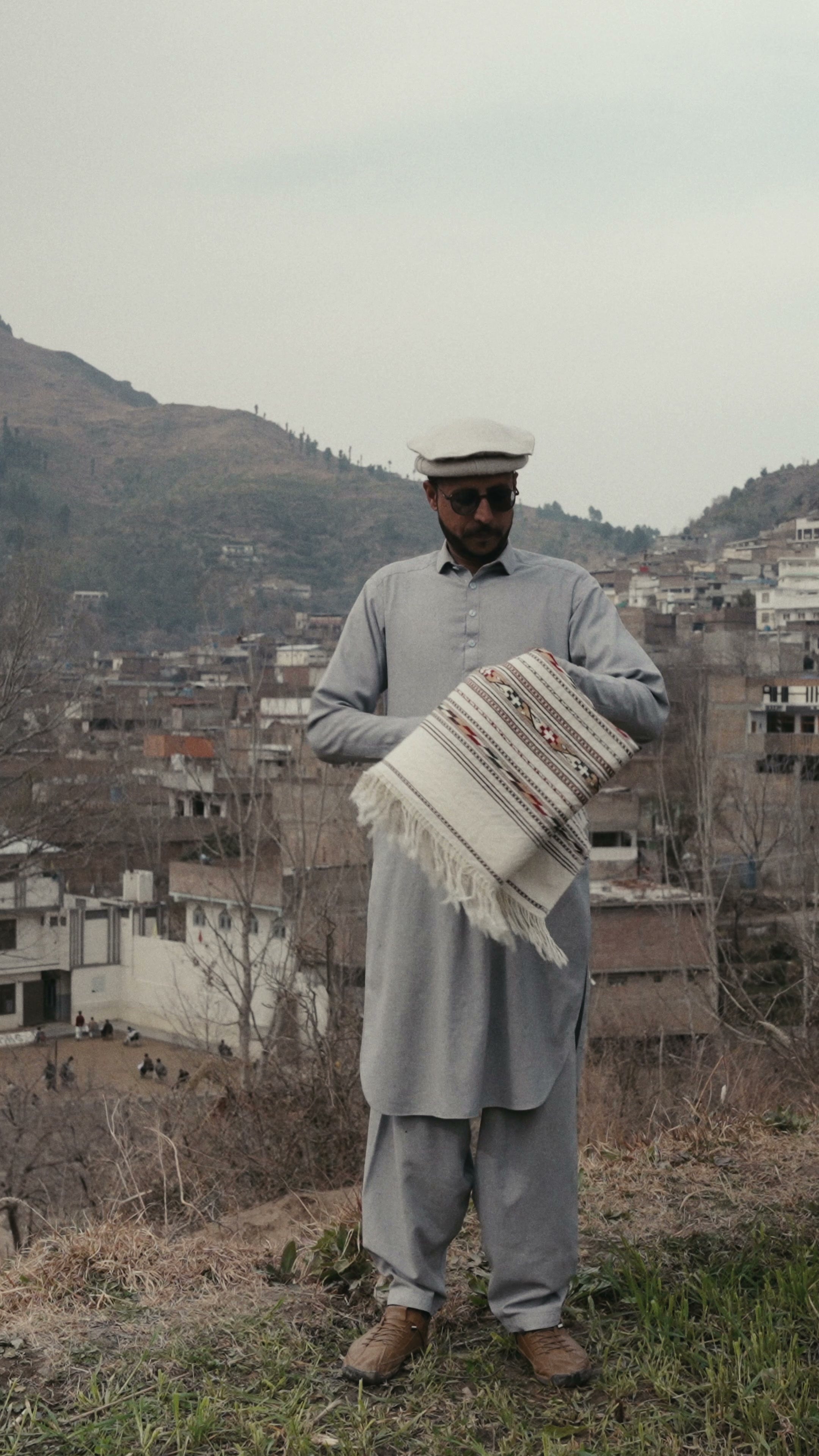 A man in a Pakistani wool cap and sunglasses holding a folded cream wool shawl with an embroidered border and fringe at his chest
