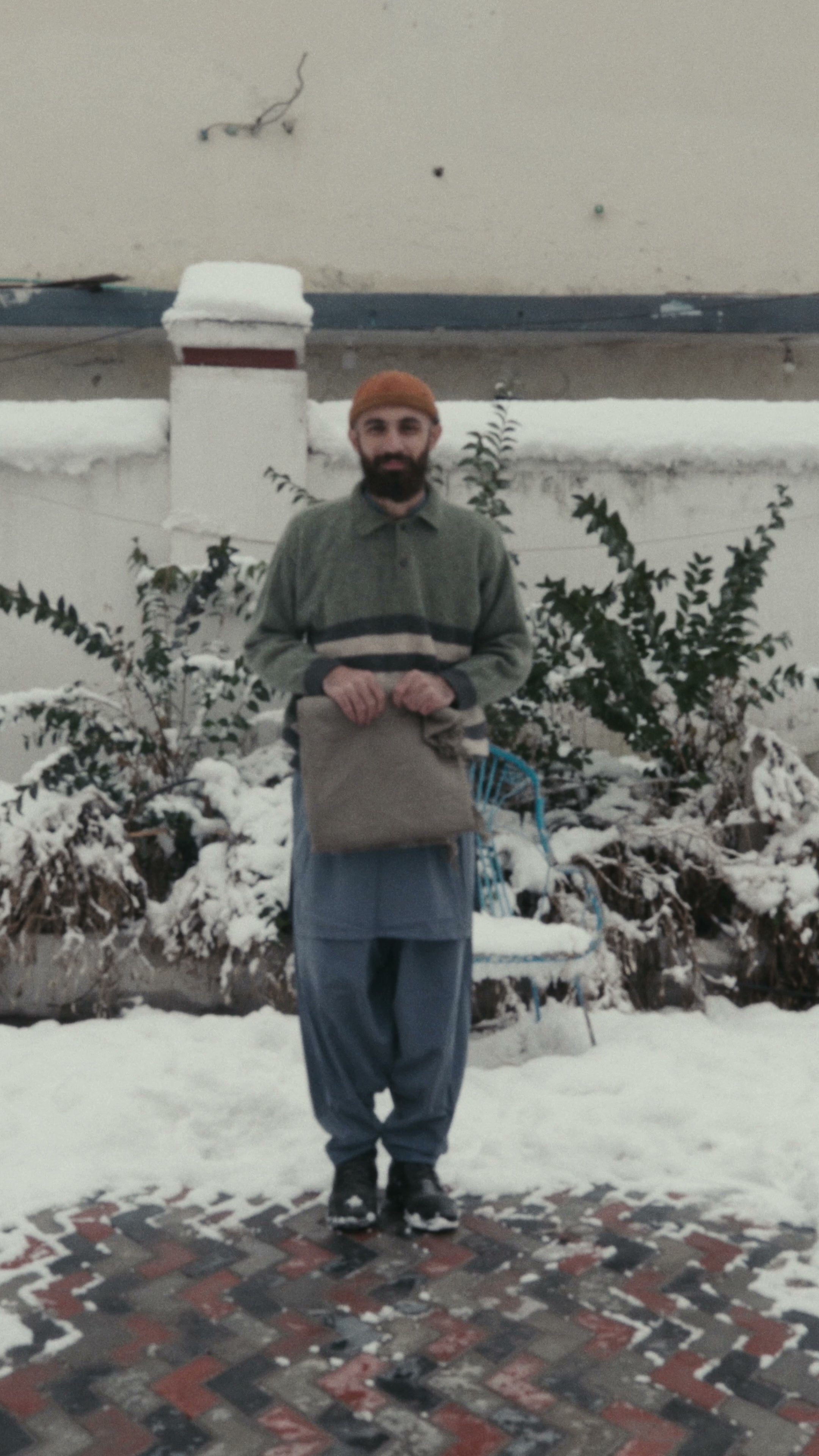 A man in a knitted cap holding a folded taupe wool shawl, standing on snow in a courtyard