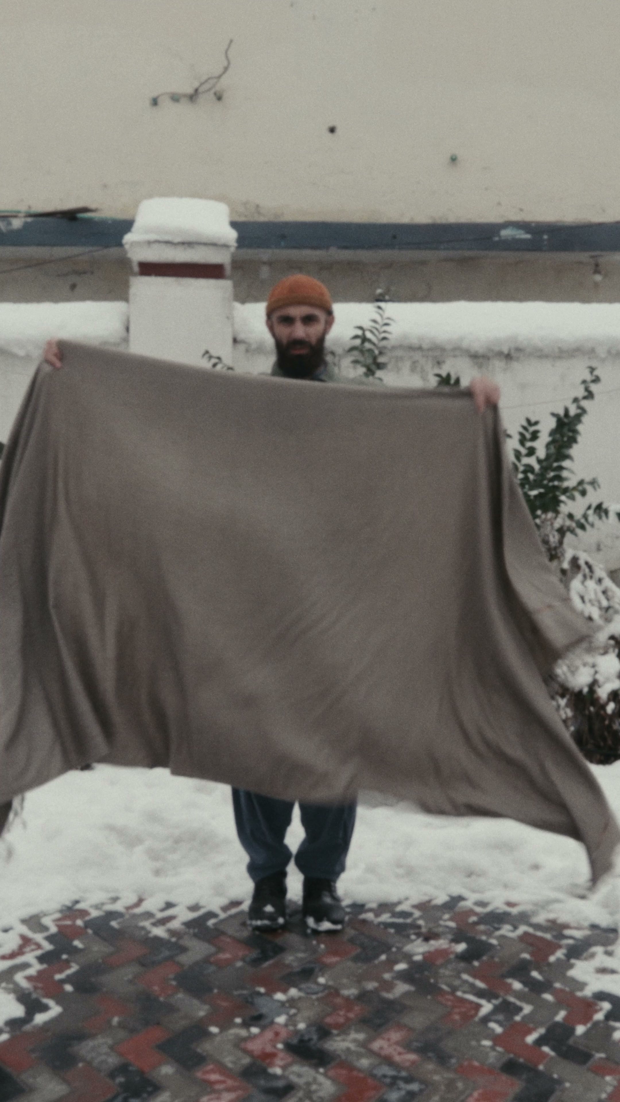 A man holding a large taupe wool shawl spread wide between his hands to show its full size, snow behind