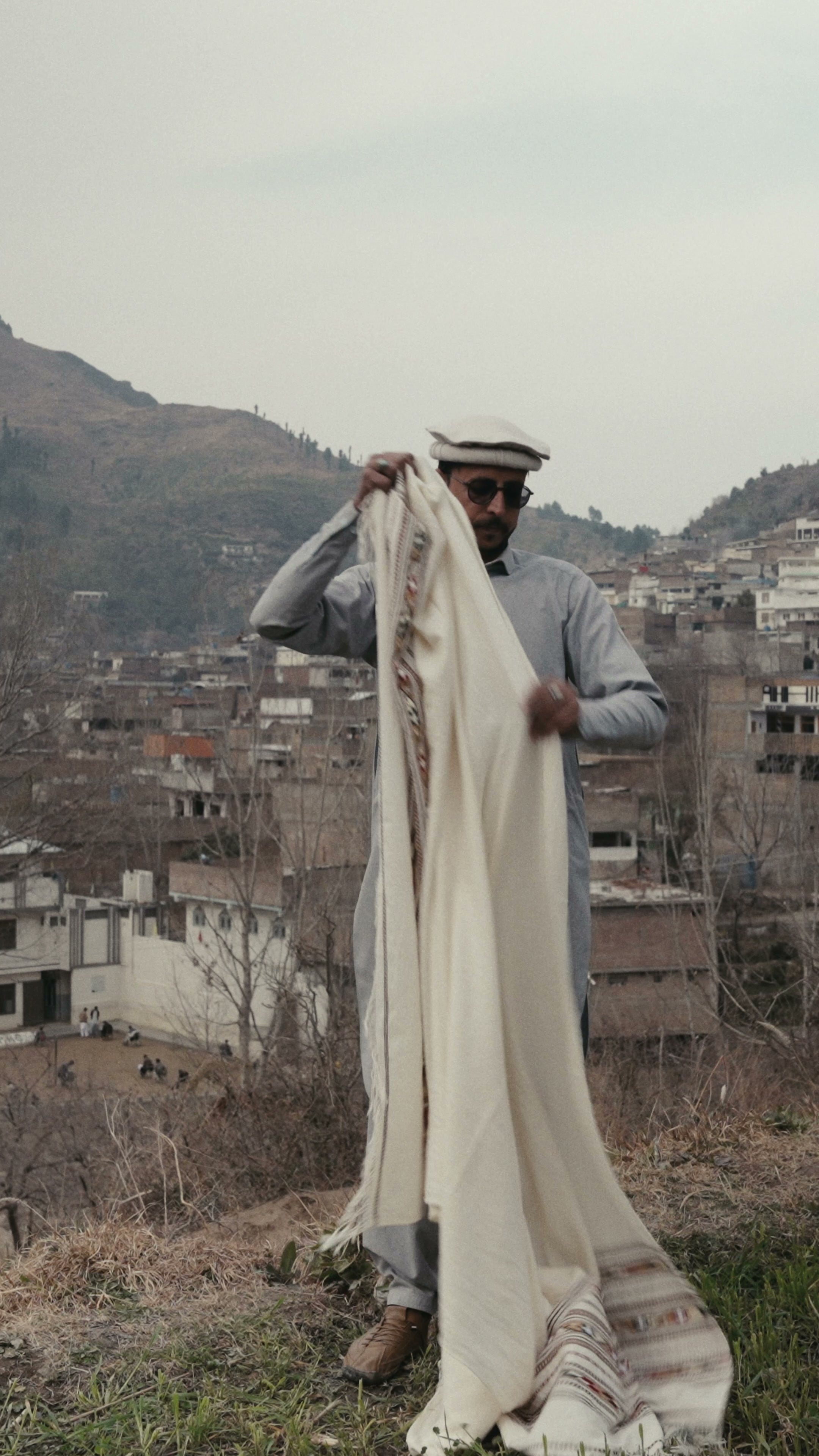 A man lifting a long cream wool shawl by one corner so it hangs full-length, about to drape it