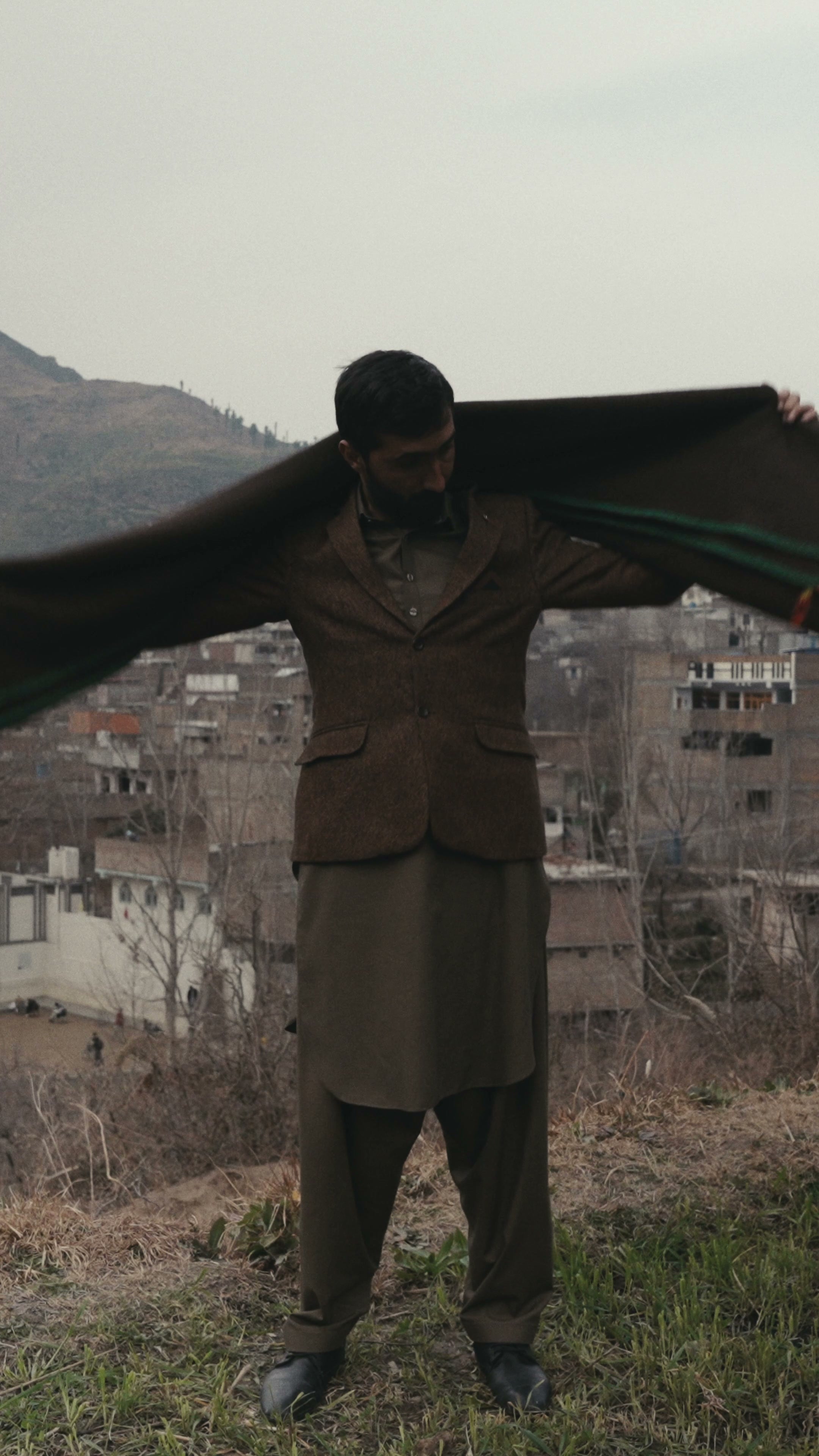 A man holding a dark brown wool shawl out with arms spread, mountains behind