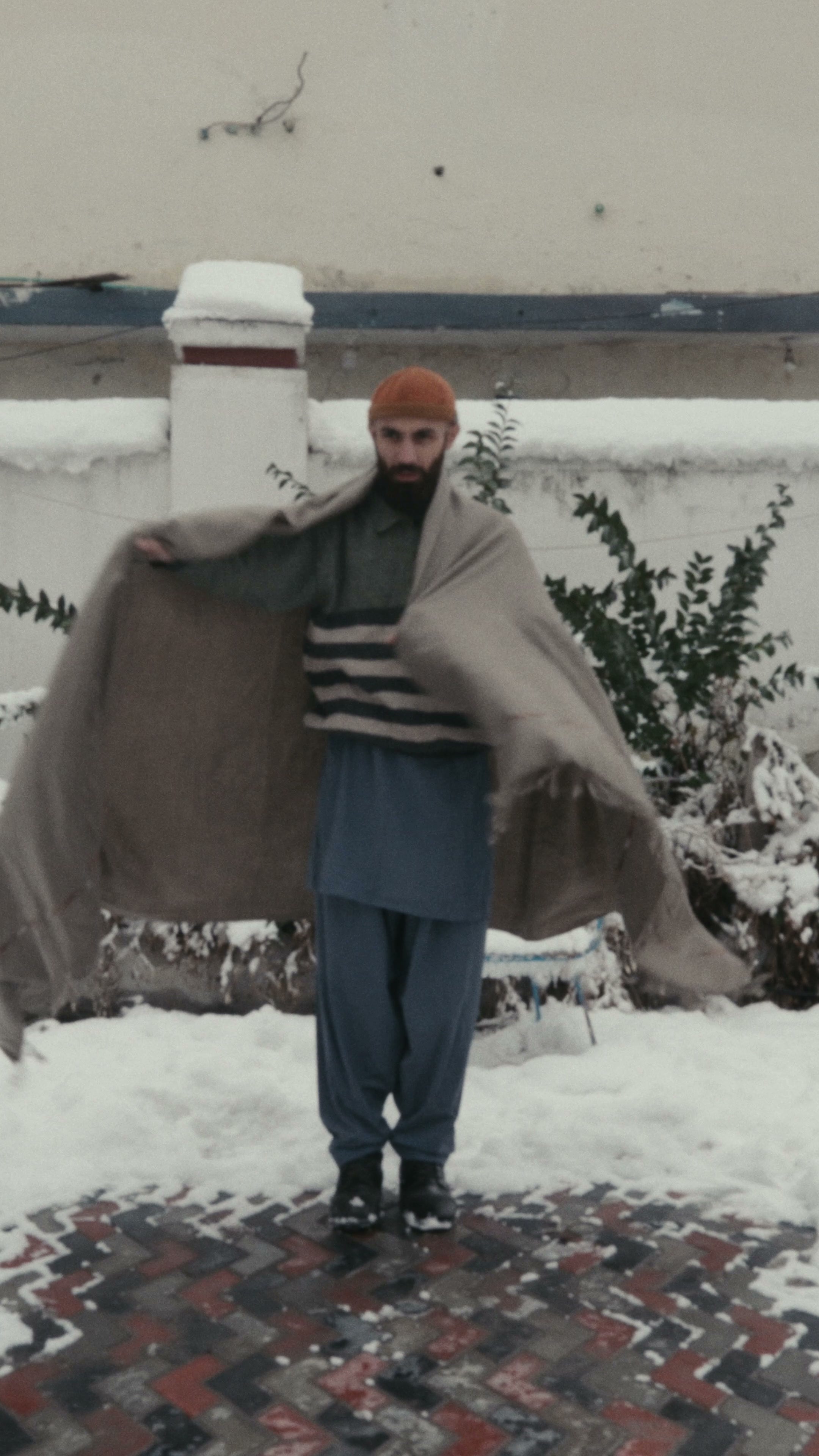 A man holding a large taupe handwoven wool shawl open across his outstretched arms, snow and bare shrubs behind
