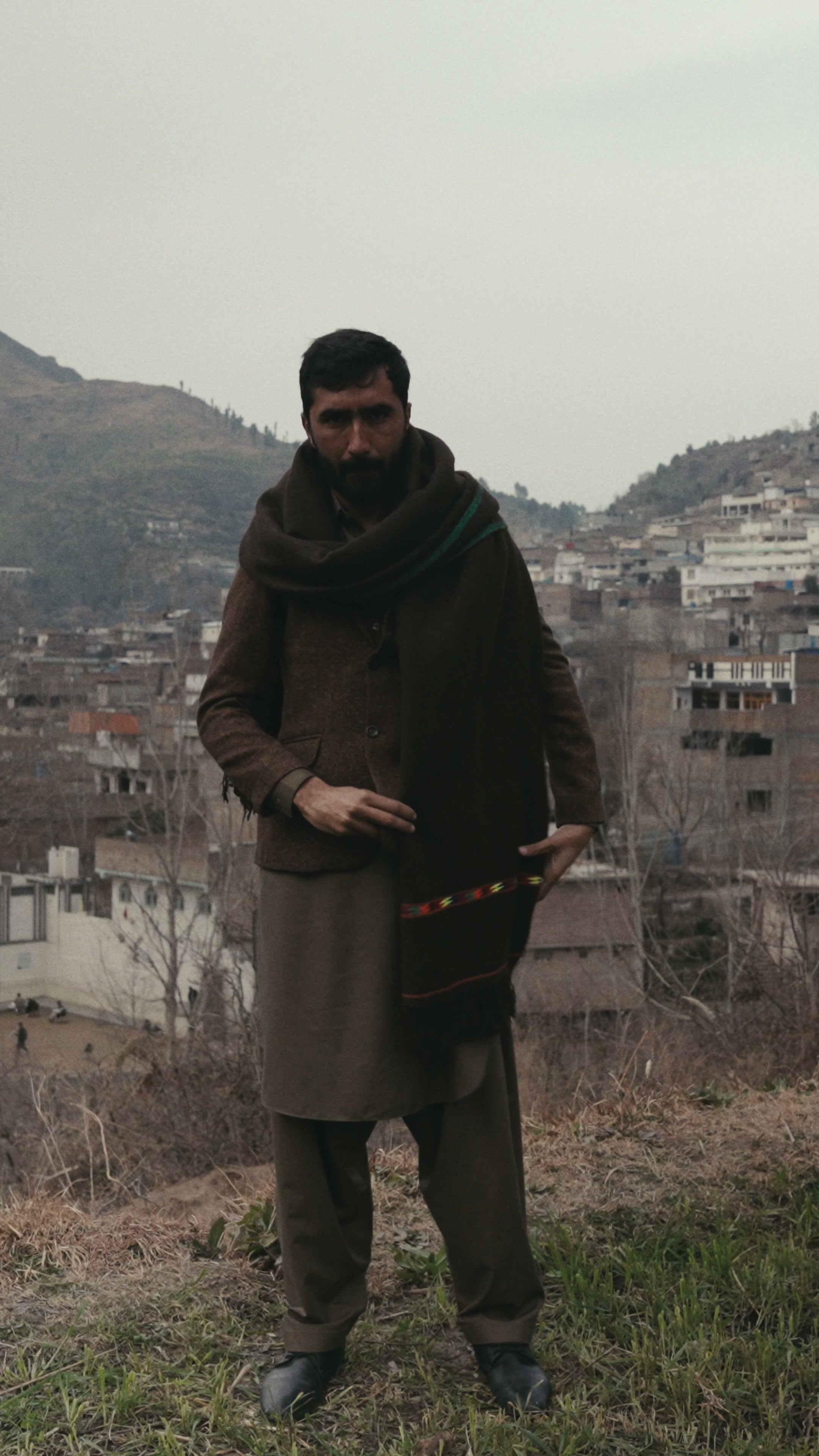 A man with a dark brown wool shawl draped over both shoulders, hands adjusting the embroidered border, a Swat town behind