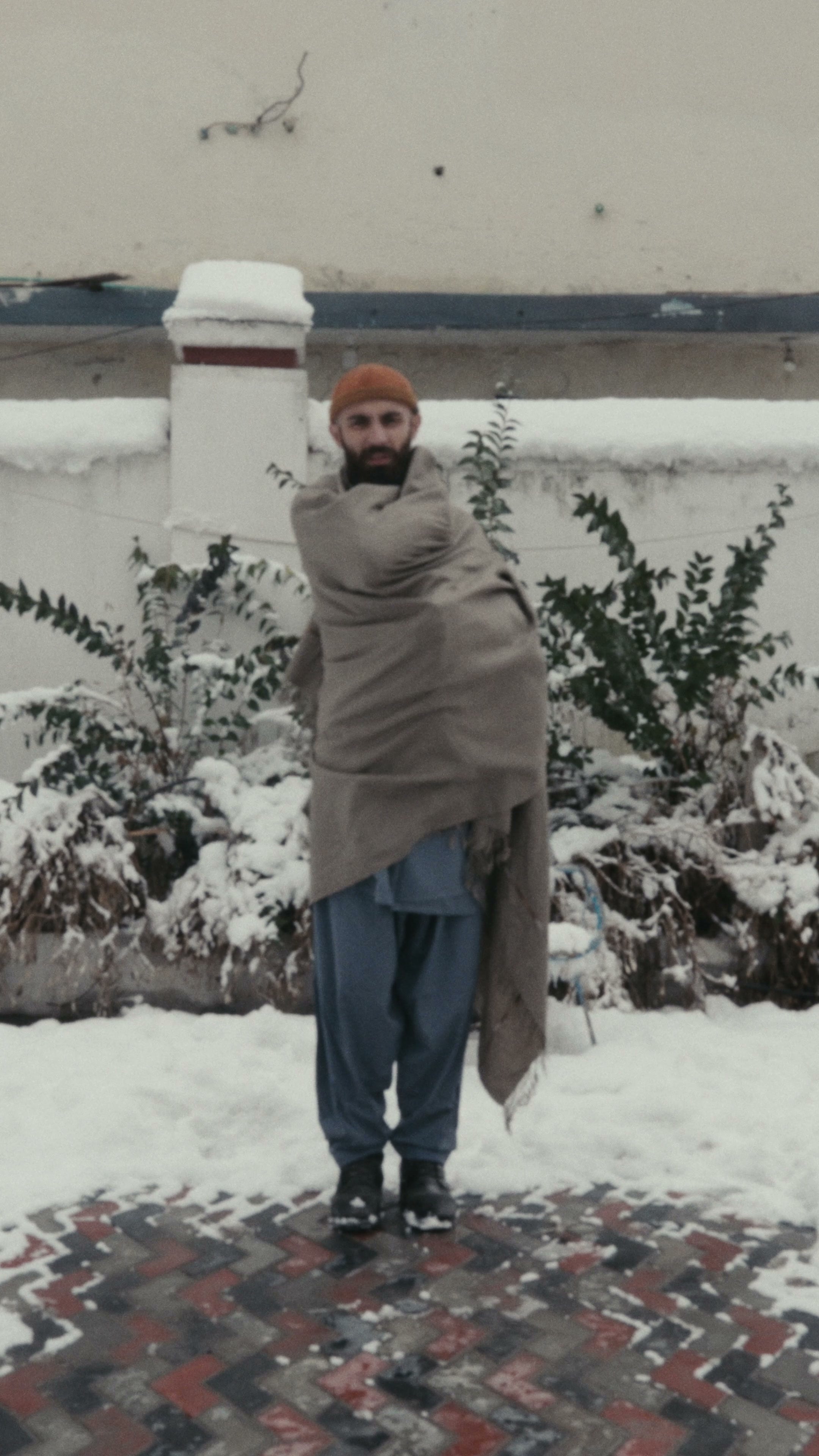 A man with a taupe handwoven wool shawl wrapped around his shoulders and body, standing on snow