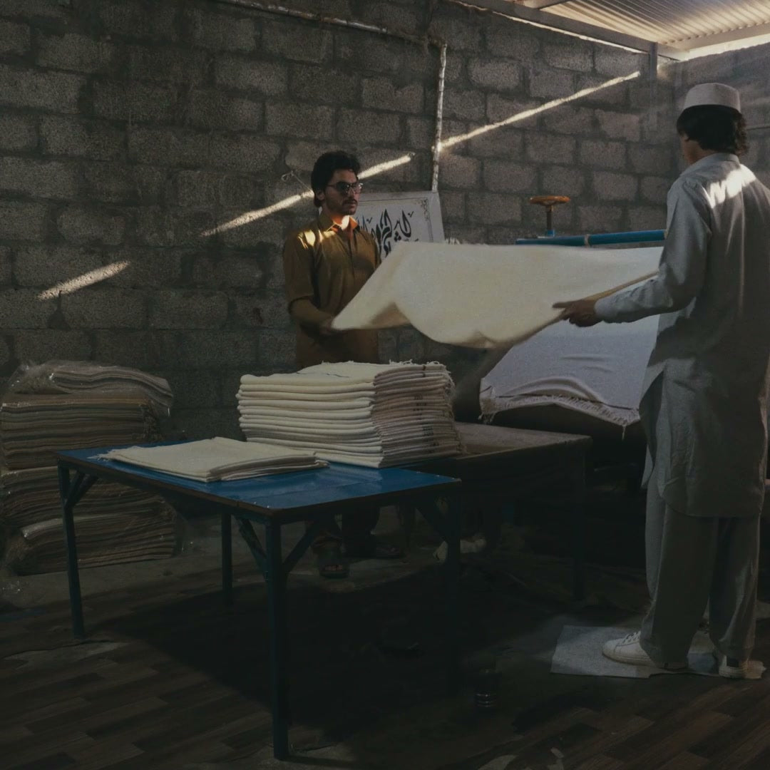Two men in a workshop folding and stacking finished cream wool shawls on a blue table