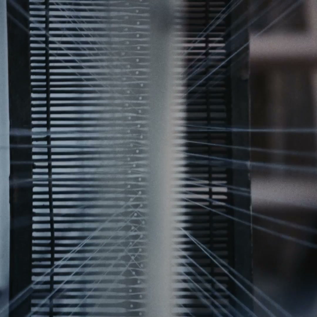 A weaver in a brown patterned sweater and blue shirt seen from behind at a wooden handloom, reed and warp visible