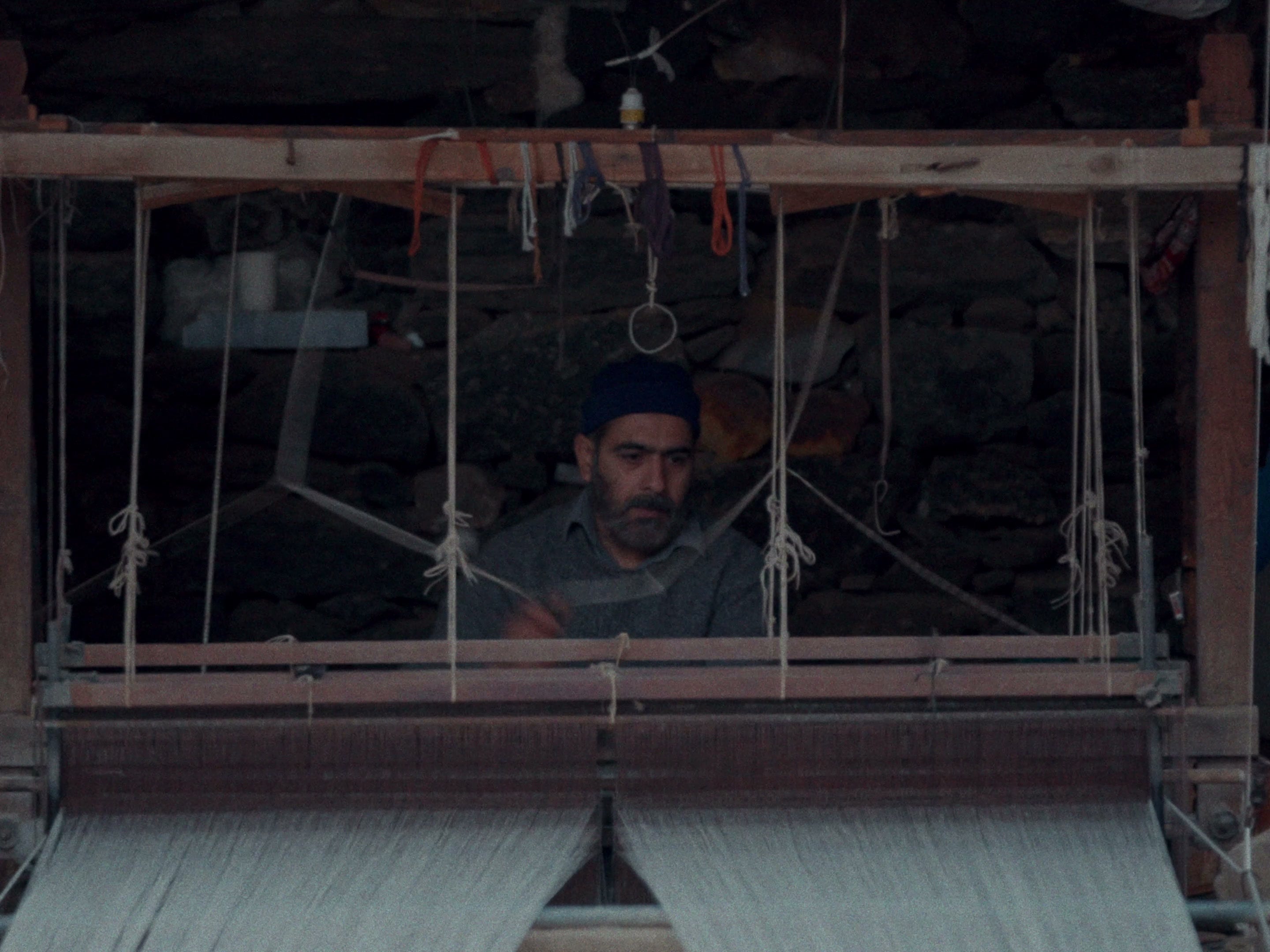 A weaver in a blue cap seated inside a large wooden handloom, working the warp, a stone wall behind
