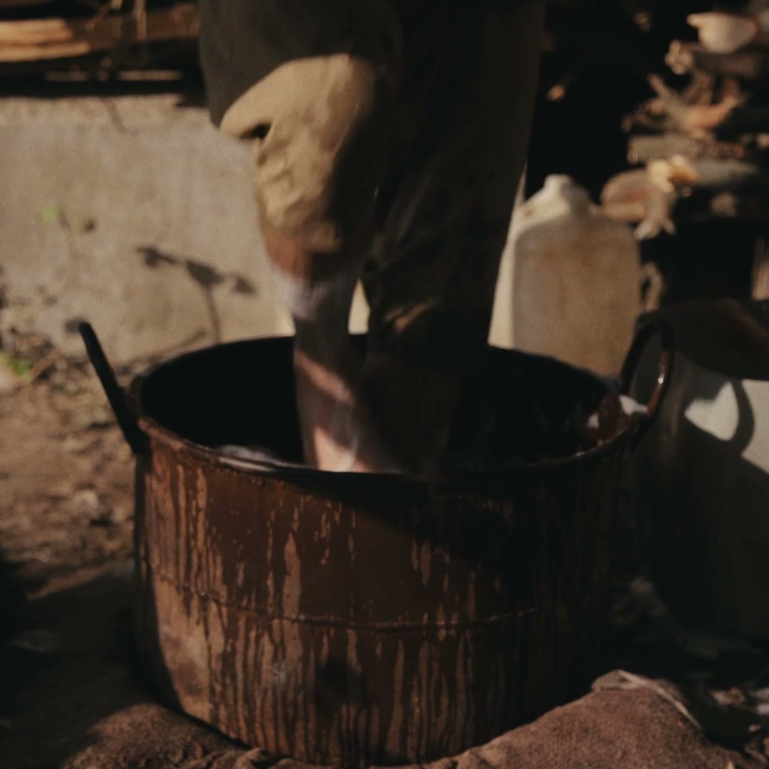 A man agitating a bundle of wool by hand in a large metal vat in a dim workshop